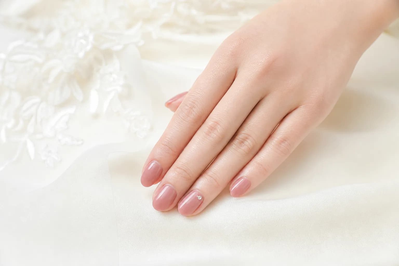 白いレースとドレス地の上の手元。くすみローズのワンカラーで、薬指だけ小さなストーンを1粒のせたブライダルネイルのデザイン例